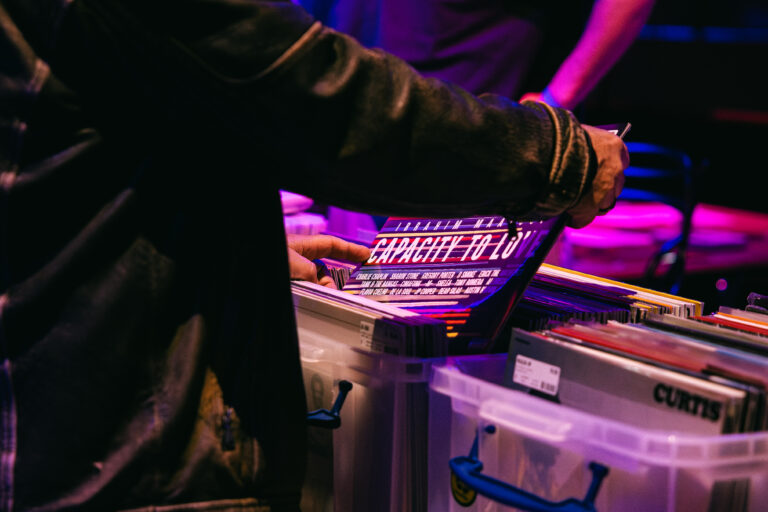 Een persoon zoekt door een doos met platen terwijl een andere persoon op de achtergrond staat.