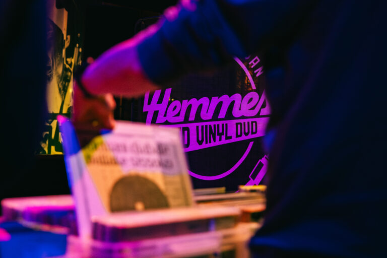 Een hand pakt een vinylplaat uit een stapel, met op de achtergrond een kleurrijk verlichte sign van een platenwinkel.