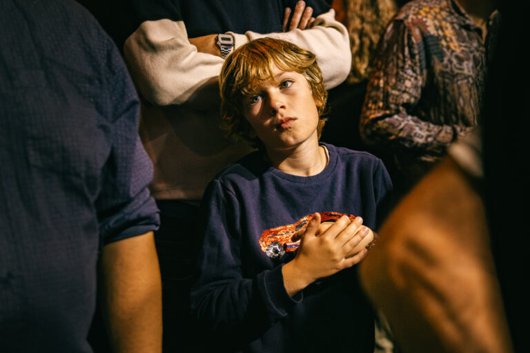 Een jongen met blond haar kijkt om zich heen terwijl hij een speelgoedauto vasthoudt, omringd door mensen.