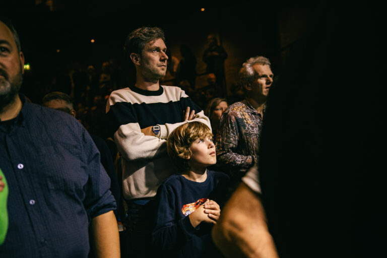 Een groep mensen, waaronder een man en een jongen, kijkt aandachtig terwijl ze naar een evenement kijken.