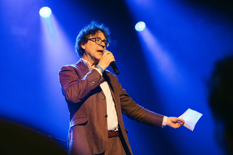 Een man in een bruine suit houdt een microfoon vast en spreekt op een podium met blauwe lichten.