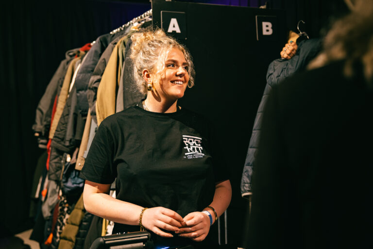 Een glimlachende jonge vrouw met een kast vol jassen op de achtergrond, terwijl ze in gesprek is.