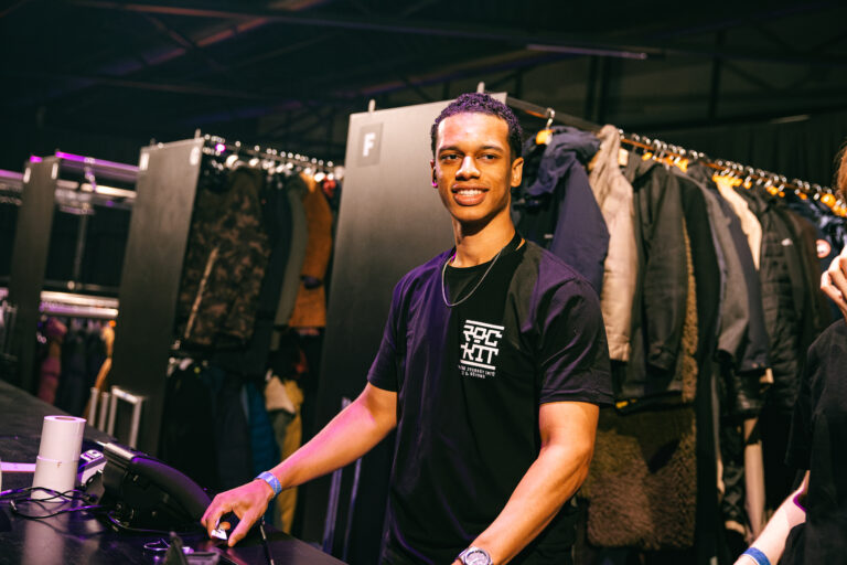 Een jonge man in een zwart T-shirt staat achter een tafel met een glimlach, terwijl kleding aan kledingrekken achter hem hangt.