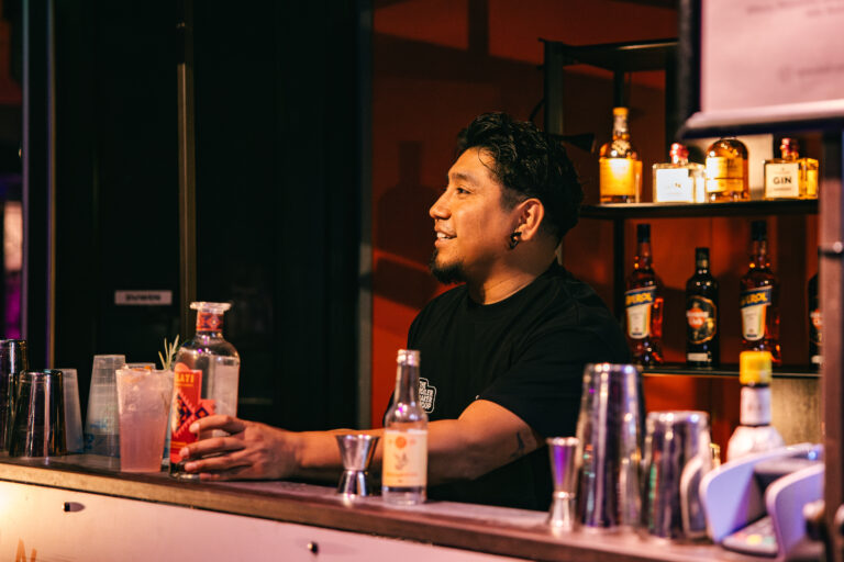 Een bartender staat achter de bar en serveert drankjes, omringd door flessen op de planken achter hem.