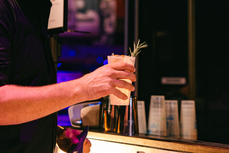 Een hand houdt een cocktail vast met een takje rozemarijn, terwijl een bar in de achtergrond zichtbaar is.