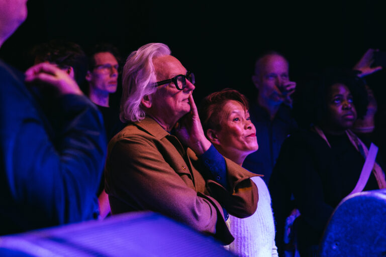Een man en een vrouw, beiden geconcentreerd en genietend, staan in een menigte tijdens een optreden met een paar andere mensen op de achtergrond.