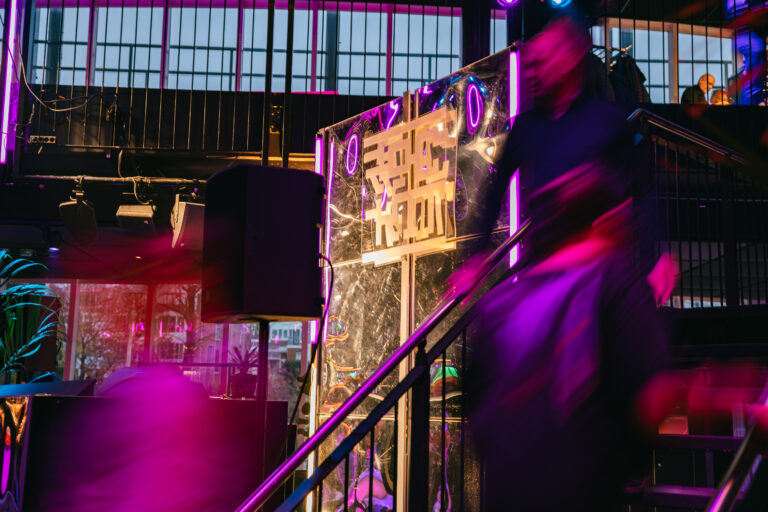 Een sfeervolle nachtclubsetting met mensen die in beweging zijn, omringd door kleurrijke verlichting en een decoratieve achterwand.