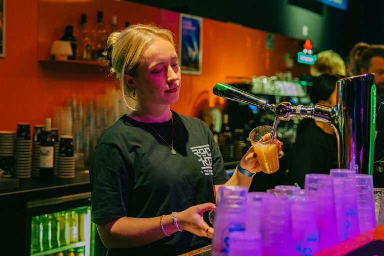 Een jonge vrouw schenkt een drankje in achter de bar, omringd door kleurrijke verlichting en glazen.