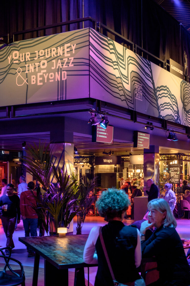 Een sfeervol interieur van een jazz- of muziekgelegenheid met een opvallend lichtgevende wandbekleding en bezoekers die met elkaar in gesprek zijn.