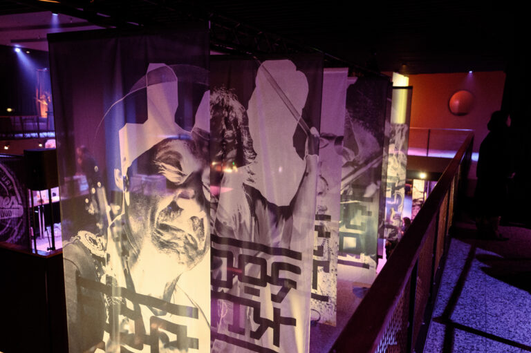 Drie grote doeken met zwart-wit afbeeldingen van mensen zijn opgezet in een sfeervolle, verlichte ruimte.