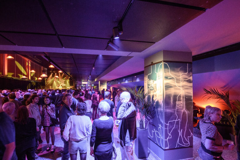 Een drukke binnenruimte met een kleurrijke verlichte bar, waar mensen met elkaar communiceren en ontspanning zoeken.