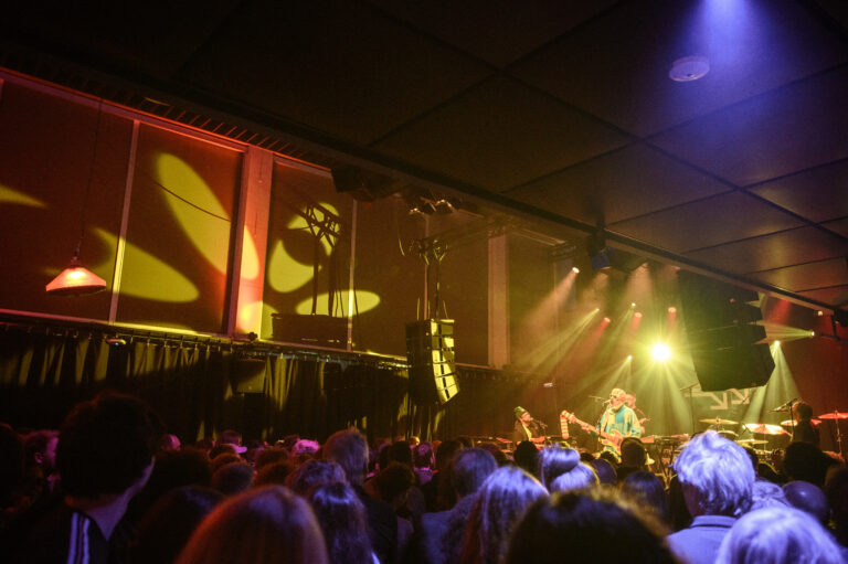 Een drukke concertzaal met een artiest op het podium, omringd door kleurrijke lichten en een enthousiast publiek.