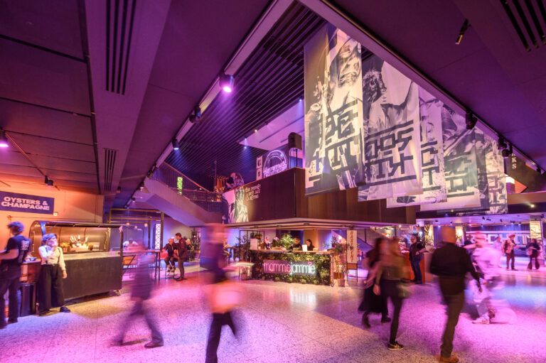 Drukke foyer van een modern gebouw met grote banners en sfeerverlichting.