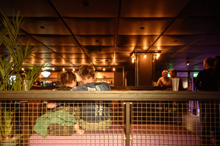 Een stel zit op een bank in een sfeervol café, omringd door andere bezoekers en sfeerverlichting.