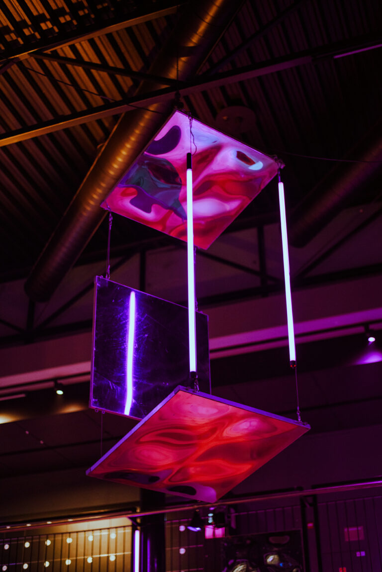 Een moderne hangende installatie met kleurrijke panelen en neonverlichting boven een sfeervolle ruimte.