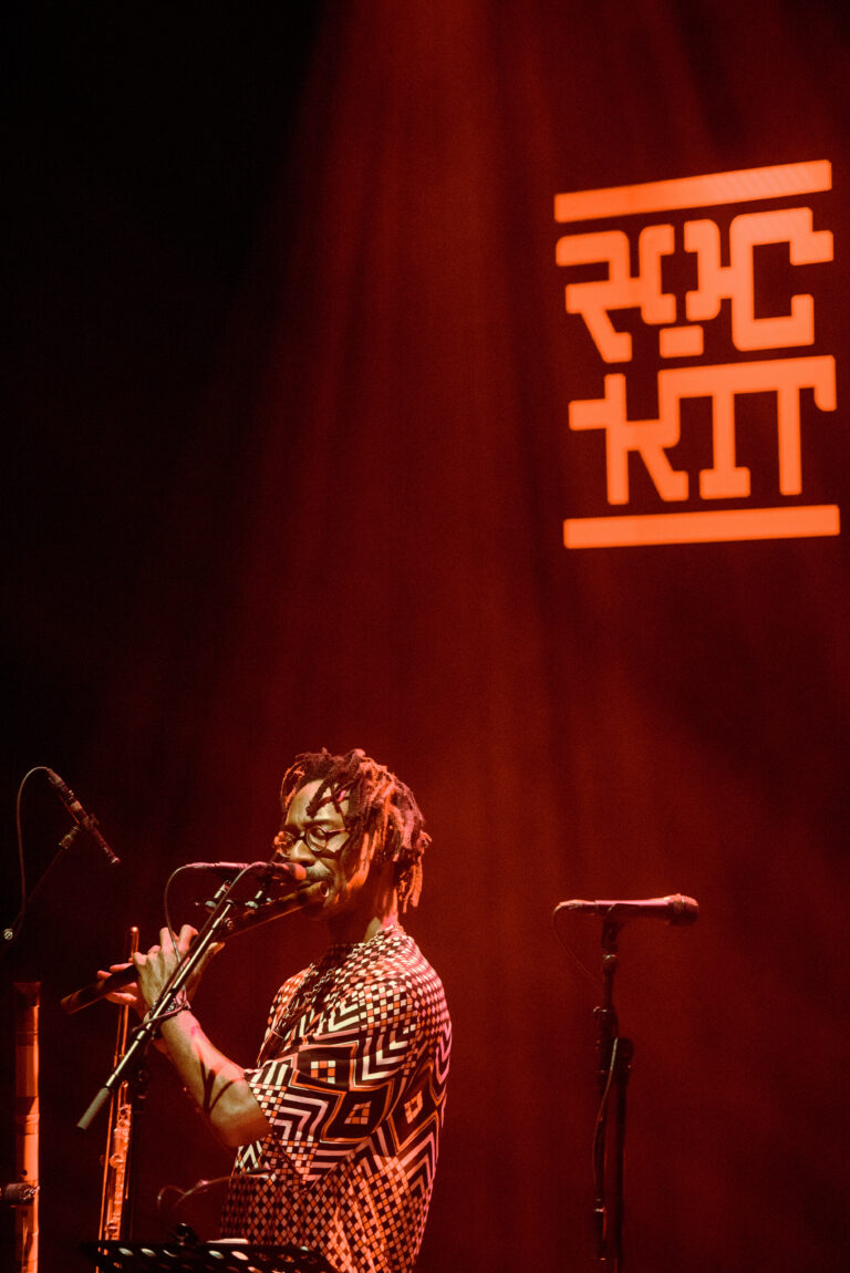 Een muzikant speelt op een podium verlicht door een rode gloed, met de naam