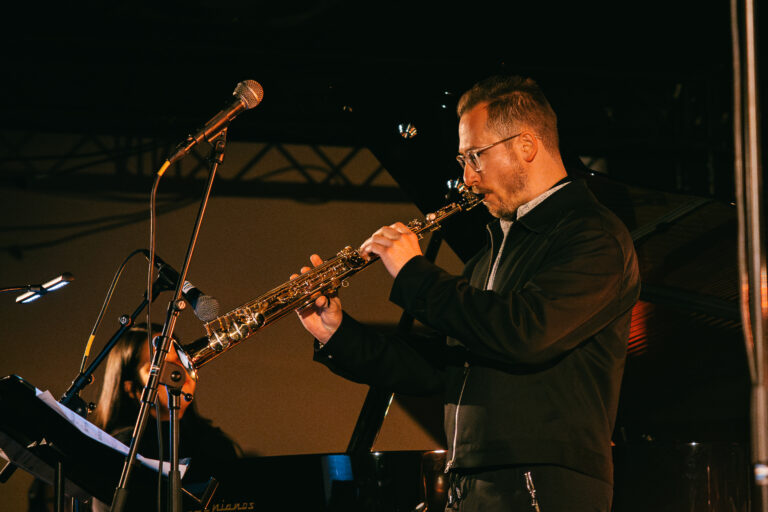 Een muzikant speelt een oboe op een podium met een microfoon en een piano op de achtergrond.