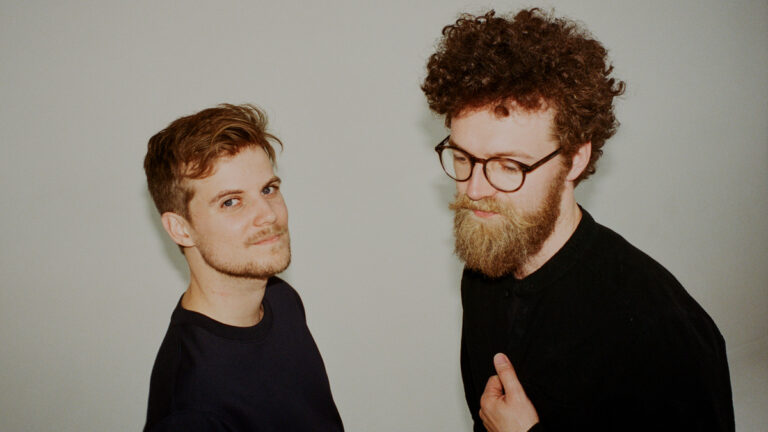 Twee mannen staan schuin naar elkaar gekeerd met een neutrale achtergrond.