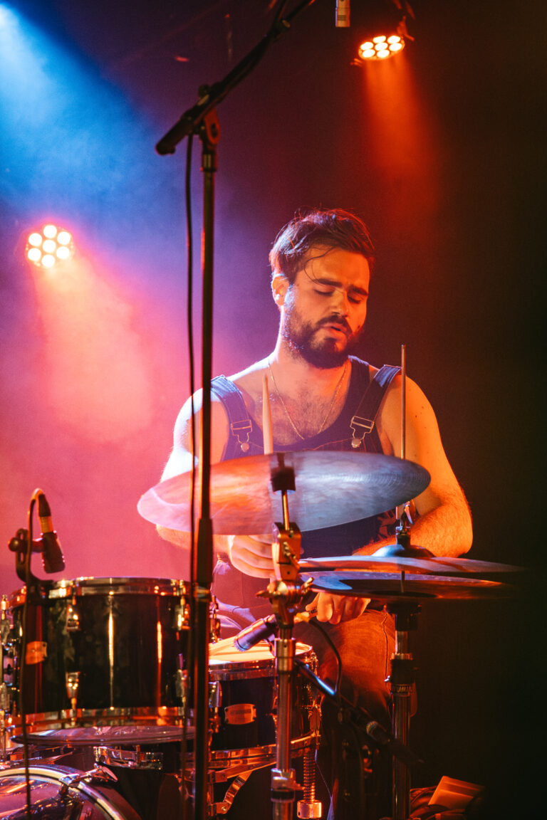 Een muzikant speelt drums op een podium, omgeven door gekleurd licht.