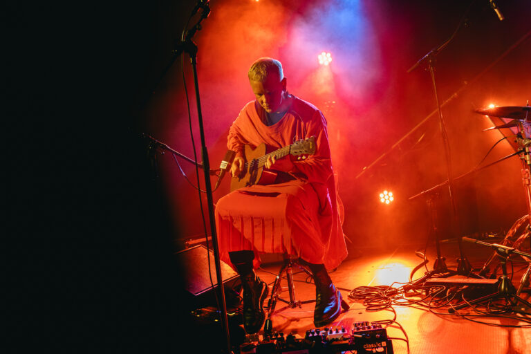 Een muzikant in een felgekleurde outfit speelt gitaar op een podium met kleurrijke lichteffecten.