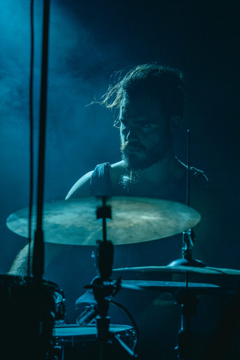 Een drummer speelt op het podium, omringd door blauwe verlichting en een lichte mist.