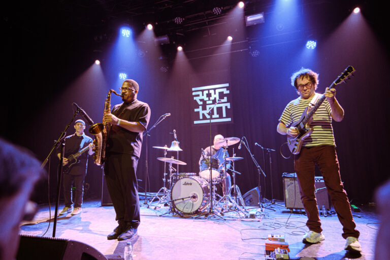 Een band speelt live op het podium, met een saxofonist, gitarist en drummer onder kleurrijke verlichting.