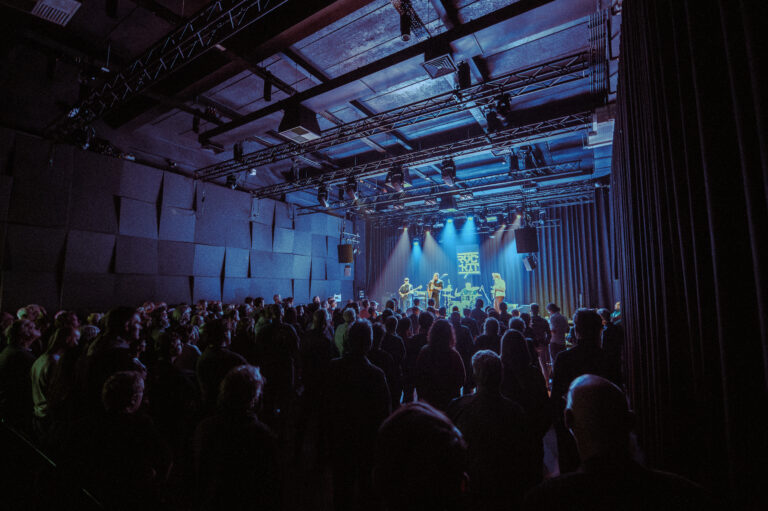 Een drukke concertzaal met een publiek dat naar een band op het podium kijkt, verlicht door blauwe lichten.