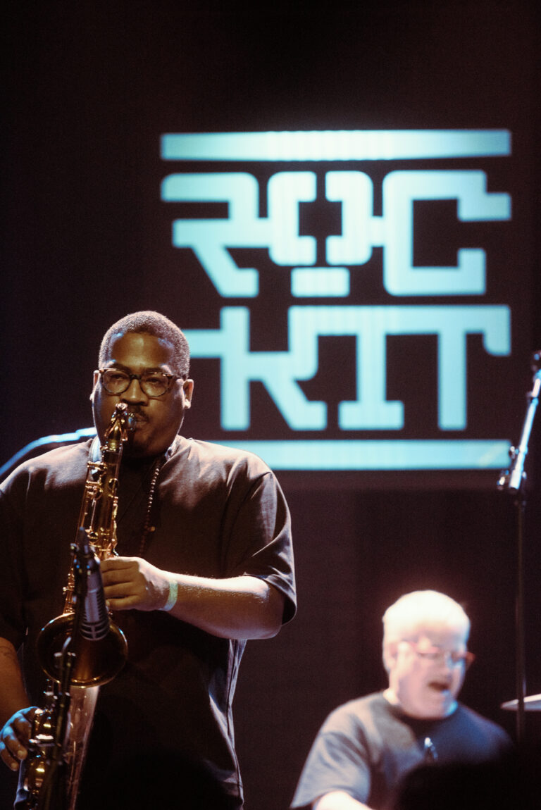 Een muzikant speelt saxofoon op het podium voor een opvallende achtergrond met het woord