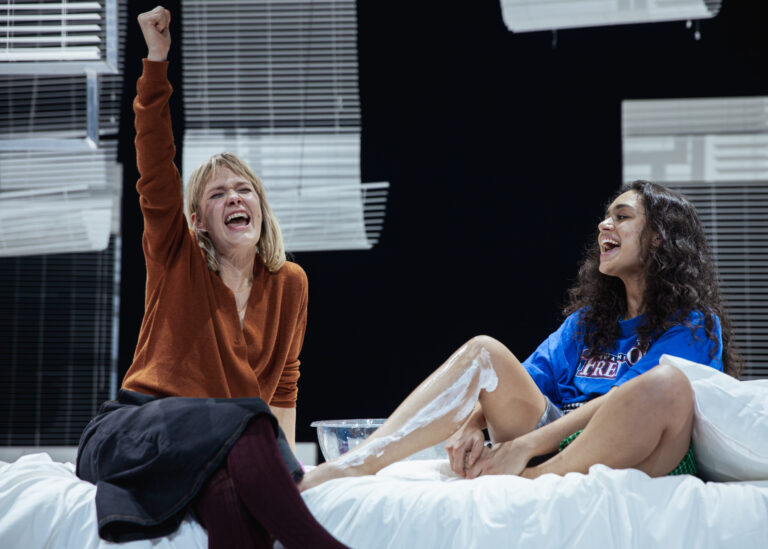 Twee lachende vrouwen zitten op een bed tijdens een theaterscène. De vrouw links, gekleed in een bruine trui, heft enthousiast een arm in de lucht. De vrouw rechts, met donker krullend haar en een blauw T-shirt, heeft een been omhoog met scheerschuim op haar scheenbeen. De achtergrond bestaat uit witte jaloezieën.