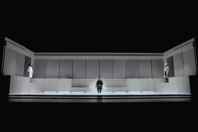 Theaterscène met drie personen op een minimalistisch toneel. Centraal zit een persoon in donkere kleding op een stoel, terwijl aan beide zijden een ander personage staat. Links staat een vrouw in een wit gewaad, rechts een persoon in een lange jas met een tas. De achtergrond bestaat uit een symmetrisch wit decor met gesloten jaloezieën.