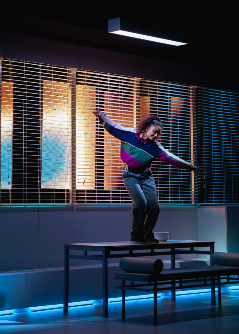 Een persoon danst met uitgespreide armen op een tafel in een fel verlicht decor. Ze dragen een kleurrijke trui in paars, groen en blauw met een grijze broek. Op de achtergrond zijn gesloten jaloezieën te zien, waar warm geel en blauw licht doorheen schijnt, wat een dynamische sfeer creëert.