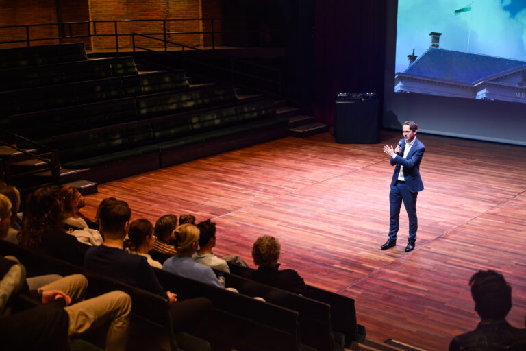 Een spreker staat op een podium in een theater voor een publiek, met een projectiescherm op de achtergrond.
