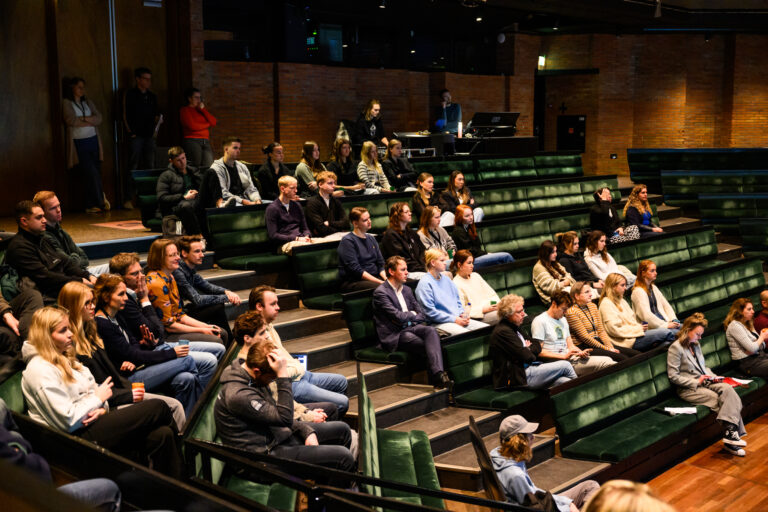 Een publiek zit op het tribune in een theater of zaal, gefocust op een presentatie of evenement.