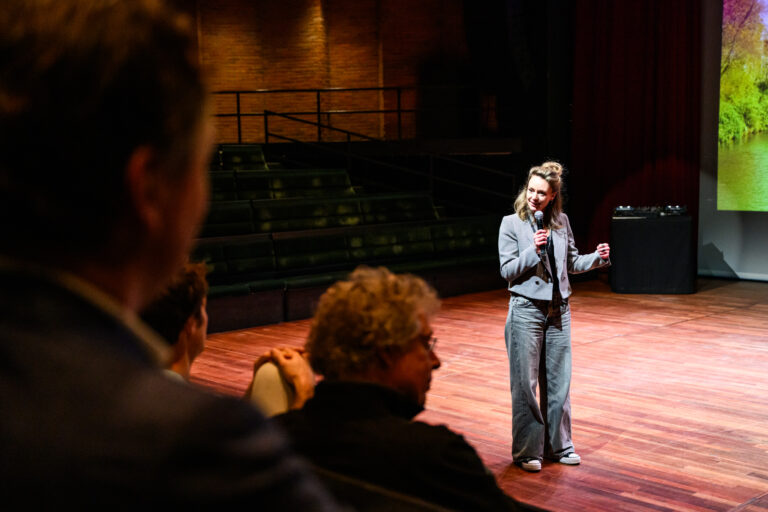 Een spreker in een grijze outfit staat op het podium en spreekt tegen het publiek in een theater.