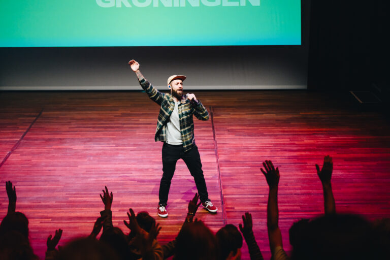 Een performer staat op een podium en steekt zijn arm in de lucht terwijl het publiek enthousiast reageert.