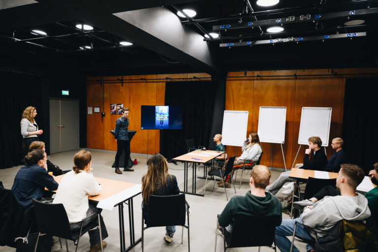 Een groepsbijeenkomst in een ruimte met houten wanden en een presentator die naar een scherm wijst, terwijl de deelnemers in een U-vormige opstelling luisteren.