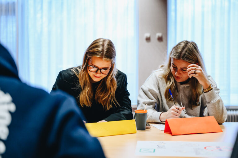 Twee vrouwen zitten aan een tafel en schrijven in notitieboekjes, met kleurrijke papieren voor zich.