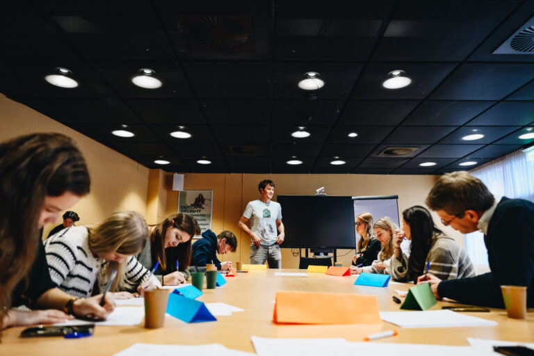 Een groep mensen in een vergaderruimte, waar iemand staat te presenteren terwijl anderen aantekeningen maken en creatief bezig zijn met papieren materialen.