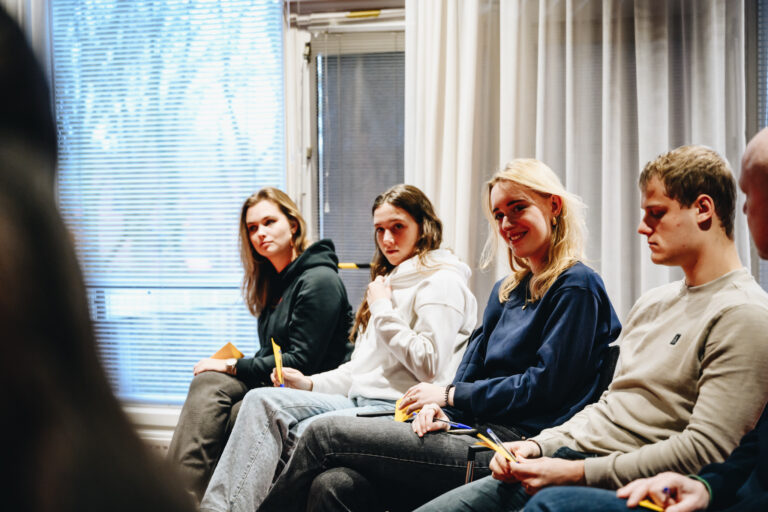 Een groep jonge mensen zit in een kamer en kijkt naar de camera, met enkele deelnemers die iets in hun handen vasthouden.
