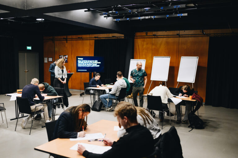 Een groep mensen in een klaslokaal werkt aan opdrachten aan verschillende tafels, terwijl instructeurs materialen presenteren op een scherm en flip-overs.