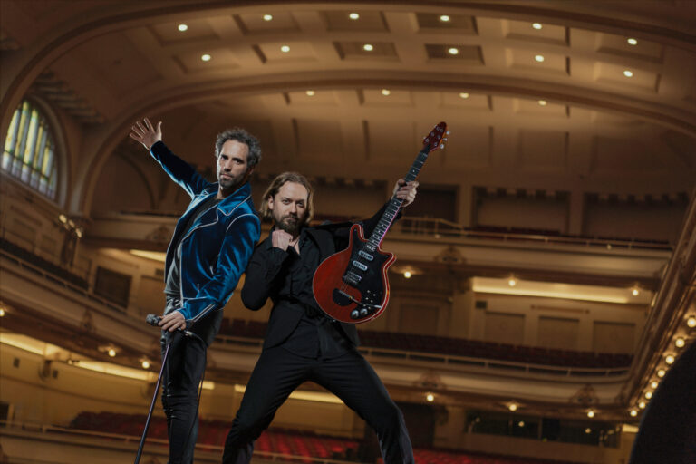 Twee muzikanten poseren op het podium van een theater, met één met een gitaar en de ander met een wandelstok.