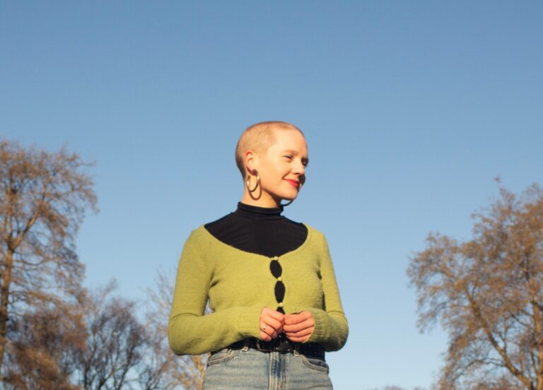 Een vrouw met een kort, helder kapsel staat buiten tegen een blauwe lucht, gedragen een groene trui en een zwarte coltrui.