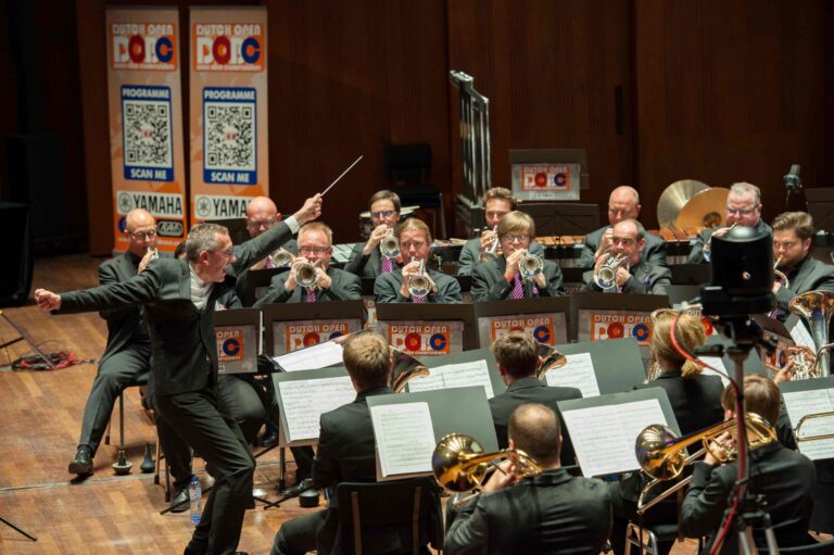 Een dirigent leidt een orkest bestaande uit muzikanten die verschillende blaasinstrumenten spelen, met een houten zaal op de achtergrond.