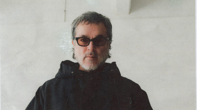 Een man met kort grijs haar, een baard, en een donkere hoodie, draagt een zonnebril en staat voor een lichte achtergrond.