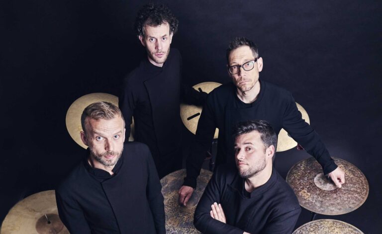 Een groep van vier muzikanten poseert voor een donkere achtergrond, met verschillende cymbalen zichtbaar.