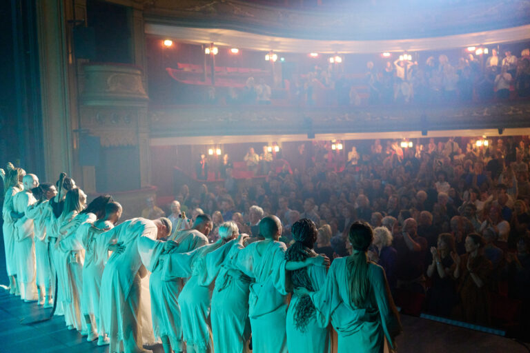 Een groep performers in witte kostuums buigt zich naar een juichend publiek in een theater.