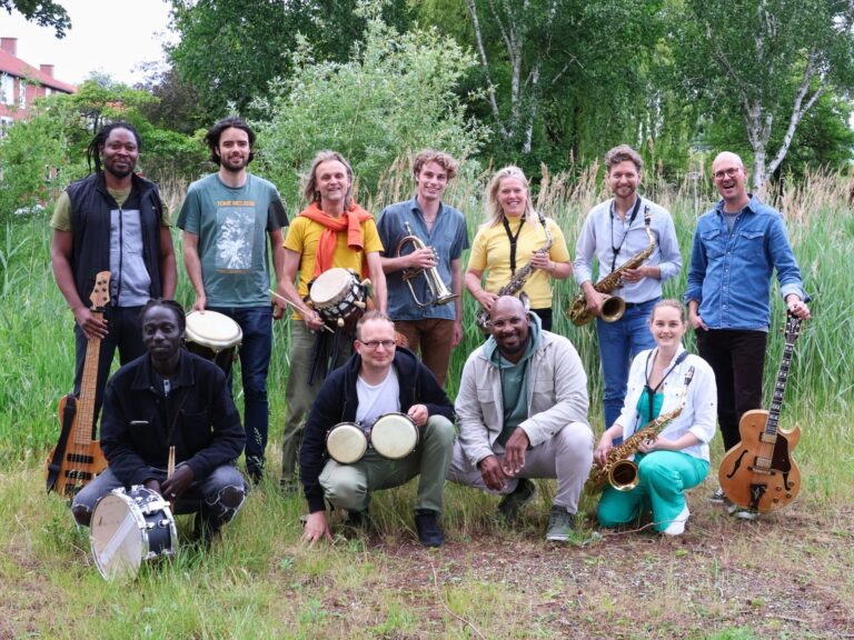 Een groep van vijftien mensen staat samen in een groene omgeving, omringd door bomen, met verschillende muziekinstrumenten in hun handen.