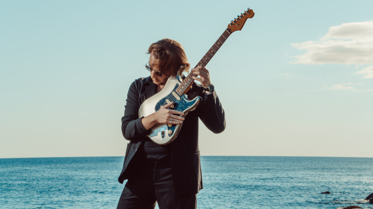 Een man in een zwart pak poseert met een elektrische gitaar voor een blauwe oceaan op een zonnige dag.