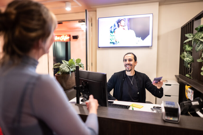 Een receptionist staat achter een balie en lacht naar een bezoeker, met een scherm op de achtergrond dat een afbeelding toont.
