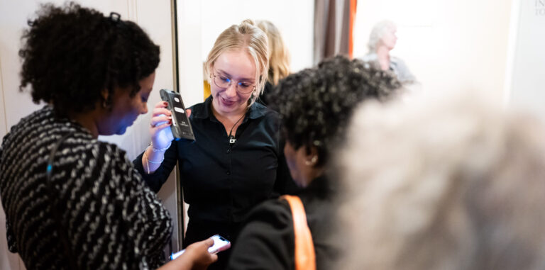 Een groep mensen staat bij een deur, waarbij één vrouw met een smartphone iets laat zien aan een andere vrouw.